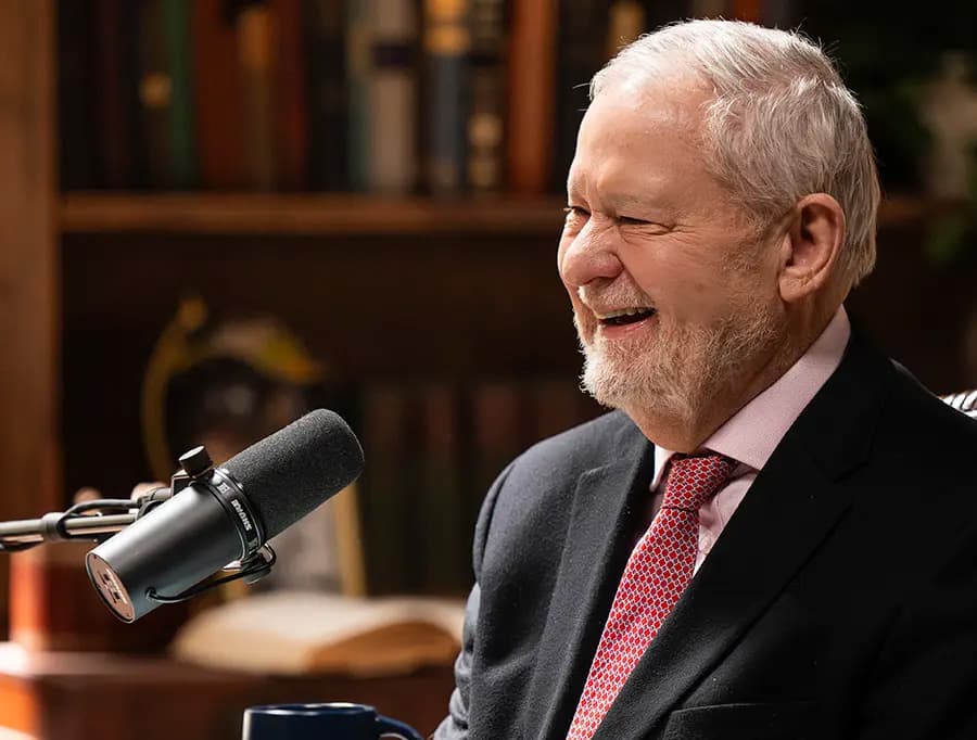 Larry Arnn laughs while recording a podcast, with a Hillsdale College mug on the table beside the microphone.