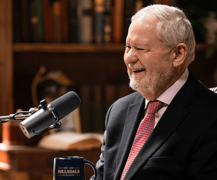 Larry Arnn laughs while recording a podcast, with a Hillsdale College mug on the table beside the microphone.